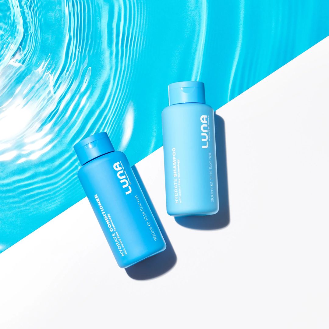 Two blue LUNA haircare bottles, one of Volume & Shine Shampoo and one of Hair Growth Serum, arranged diagonally on a split white and bright blue water-textured background.
