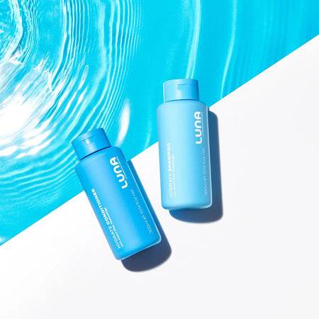 Two blue LUNA haircare bottles, one of Volume & Shine Shampoo and one of Hair Growth Serum, arranged diagonally on a split white and bright blue water-textured background.