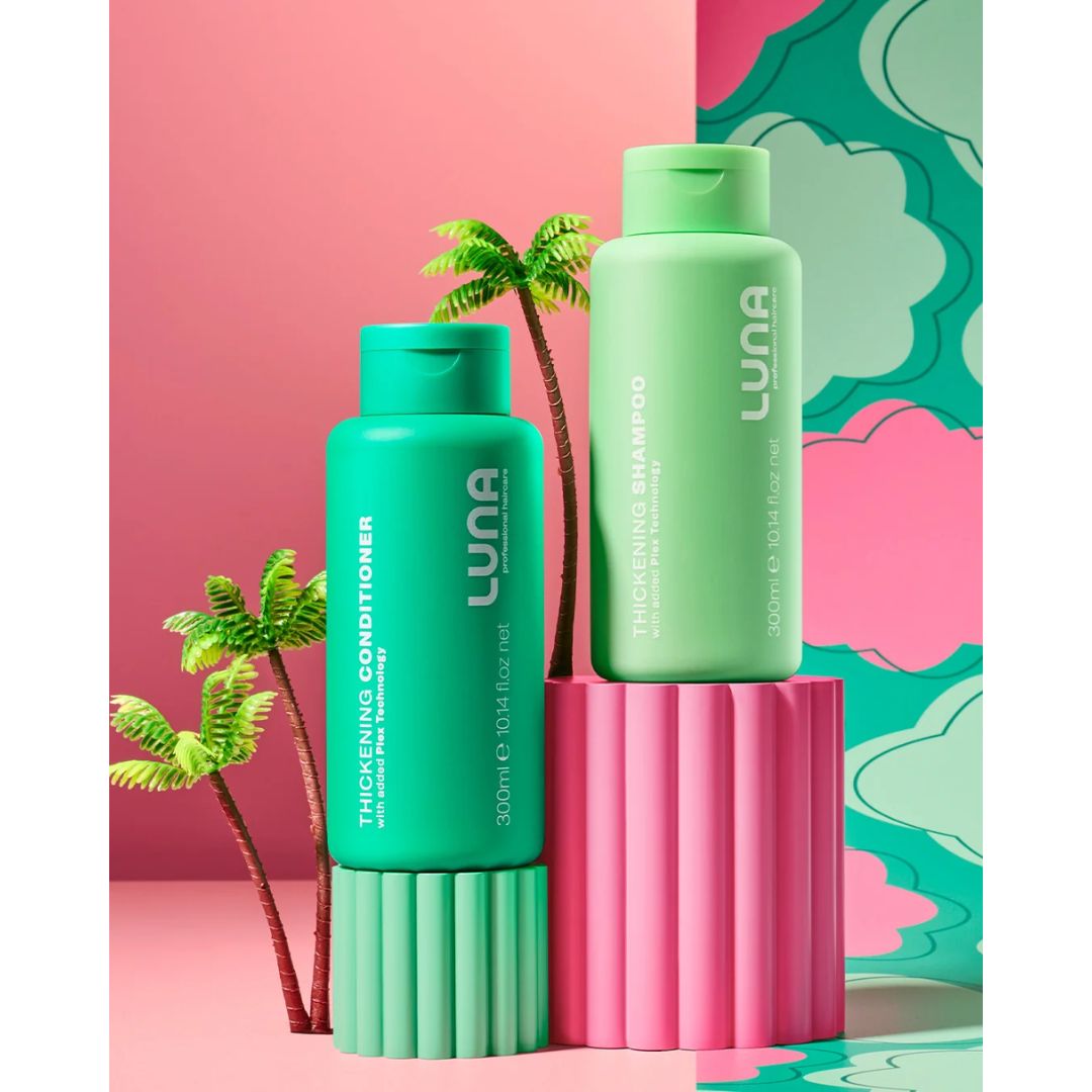 Two LUNA haircare bottles, a thickening conditioner in teal and a thickening shampoo in mint green, displayed on pink and mint green pedestal blocks against a pink and teal patterned background with small palm tree props.