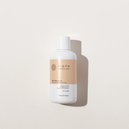 A 100ml bottle of mane combinator hair hair oil, featuring a white bottle with a beige label, set against a warm off-white background with a soft shadow.