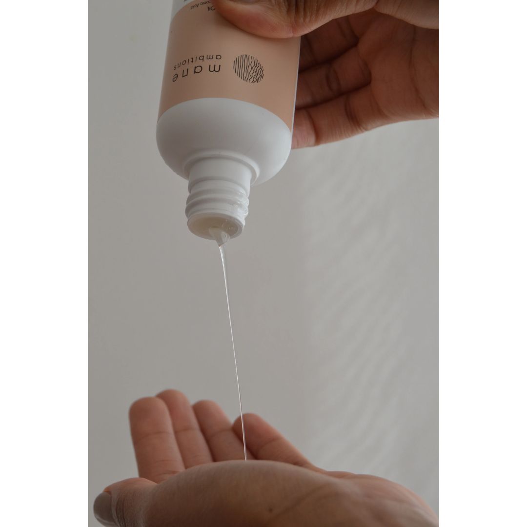 A hand holding a beige and white bottle of hair conditioner, with a thin stream of clear conditioner being poured into an open palm below.