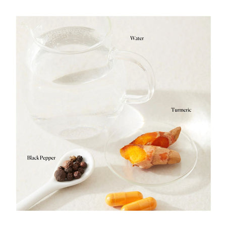 A clear glass pitcher filled with water on a light background, with labeled ingredients nearby: turmeric root on a small glass dish, black pepper in a white spoon, and two orange capsules. Text labels read Water, Turmeric, and Black Pepper.