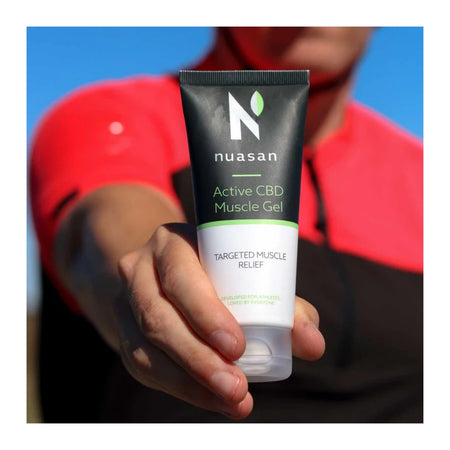 A hand holding a squeeze tube of Nuasan Active CBD Muscle Gel against a blurred background of a person wearing a red shirt and blue sky.