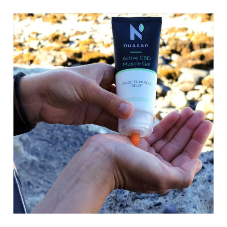 A person's hands holding and squeezing a tube of Nuasan Active CBD Muscle Gel, with gel dripping into an open palm, set against a blurred outdoor rocky background.
