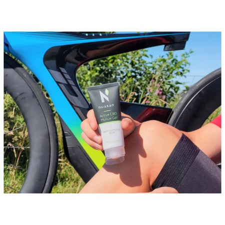 A hand holding a squeeze tube of nuxan Active CBD Muscle Gel in front of a bright blue bicycle frame outdoors.