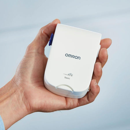 A hand holding an Omron compact AFR basic monitor device, which is white with a blue button, against a soft light blue background.