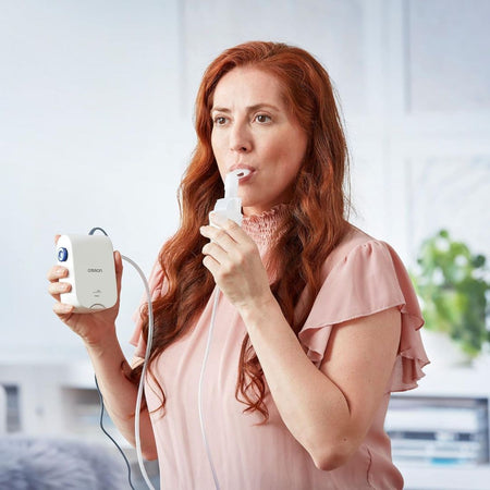 A woman with long red hair wearing a light pink top uses a Respiron nebulizer, holding the white device in one hand and the mouthpiece in the other, against a bright, softly blurred indoor background.
