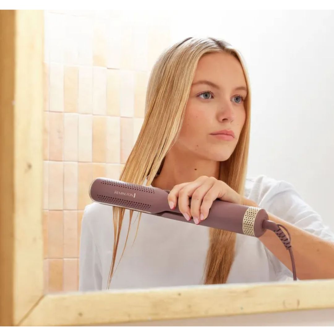A woman with long blonde hair uses a brown hair straightener in front of a mirror, showing the product in use in a bathroom setting.