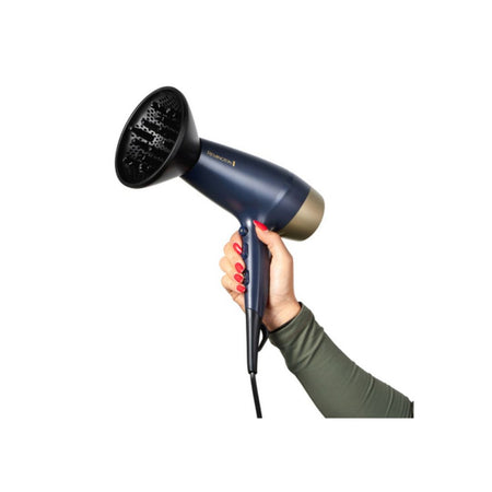 A hand with red nail polish holds a navy blue and gold hair dryer with a round diffuser attachment against a solid white background.