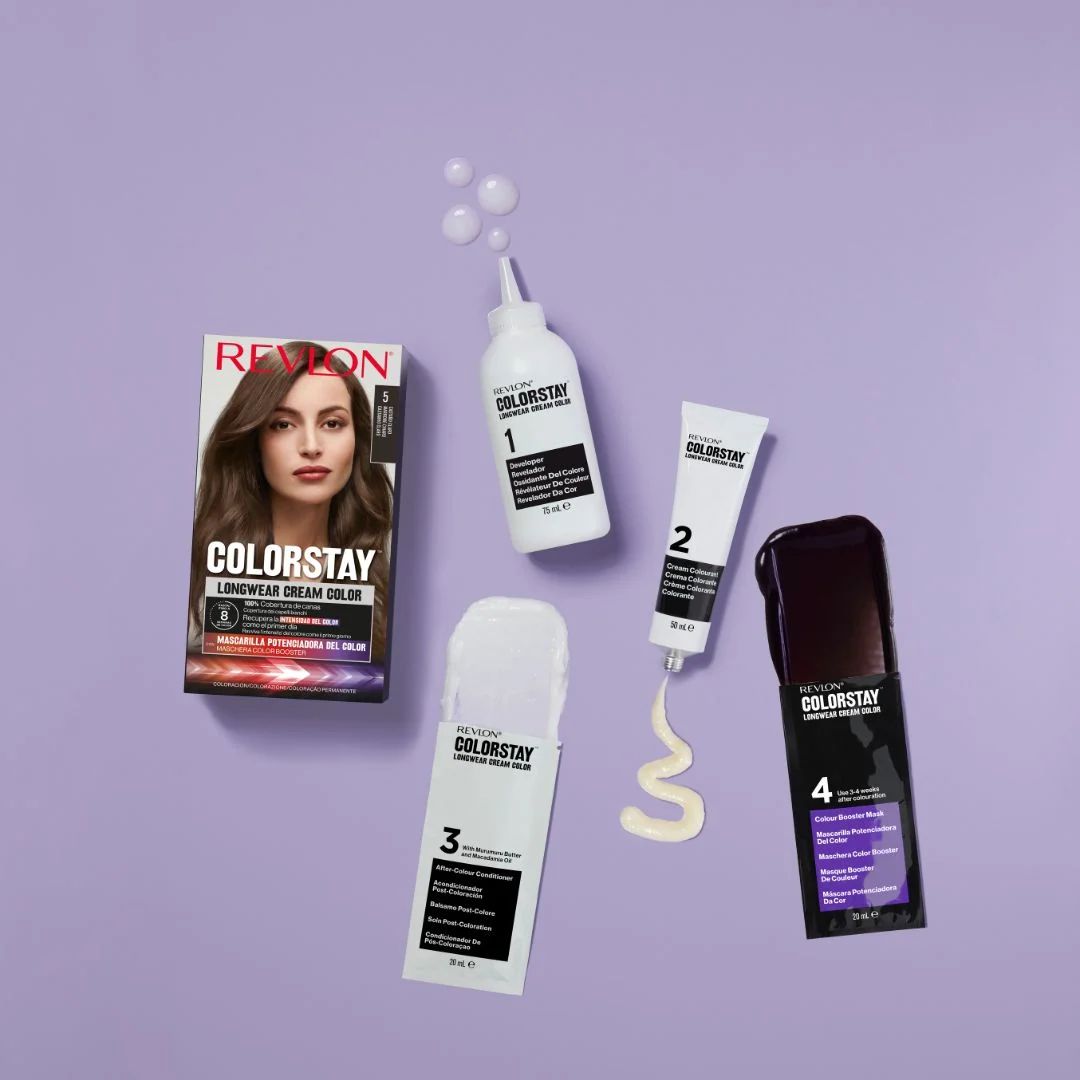 Revlon ColorStay longwear cream hair color kit with four components arranged on a solid lavender background: a hair color box, a white bottle of developer, a white tube of cream, a white cap, and a black packet with purple text.