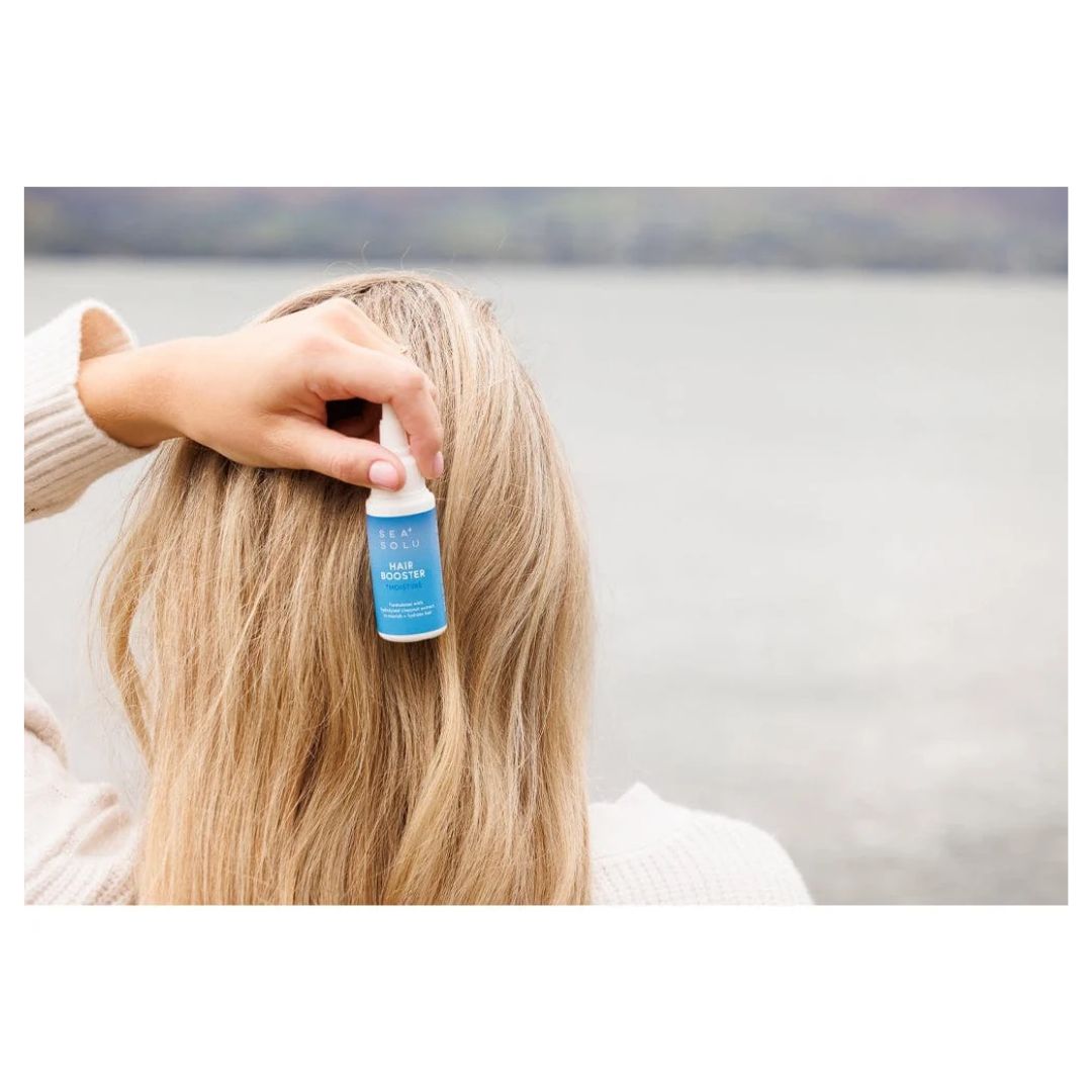 A person with long blonde hair seen from behind outdoors near water, holding a small blue spray bottle against their hair. The person wears a cream knit top.