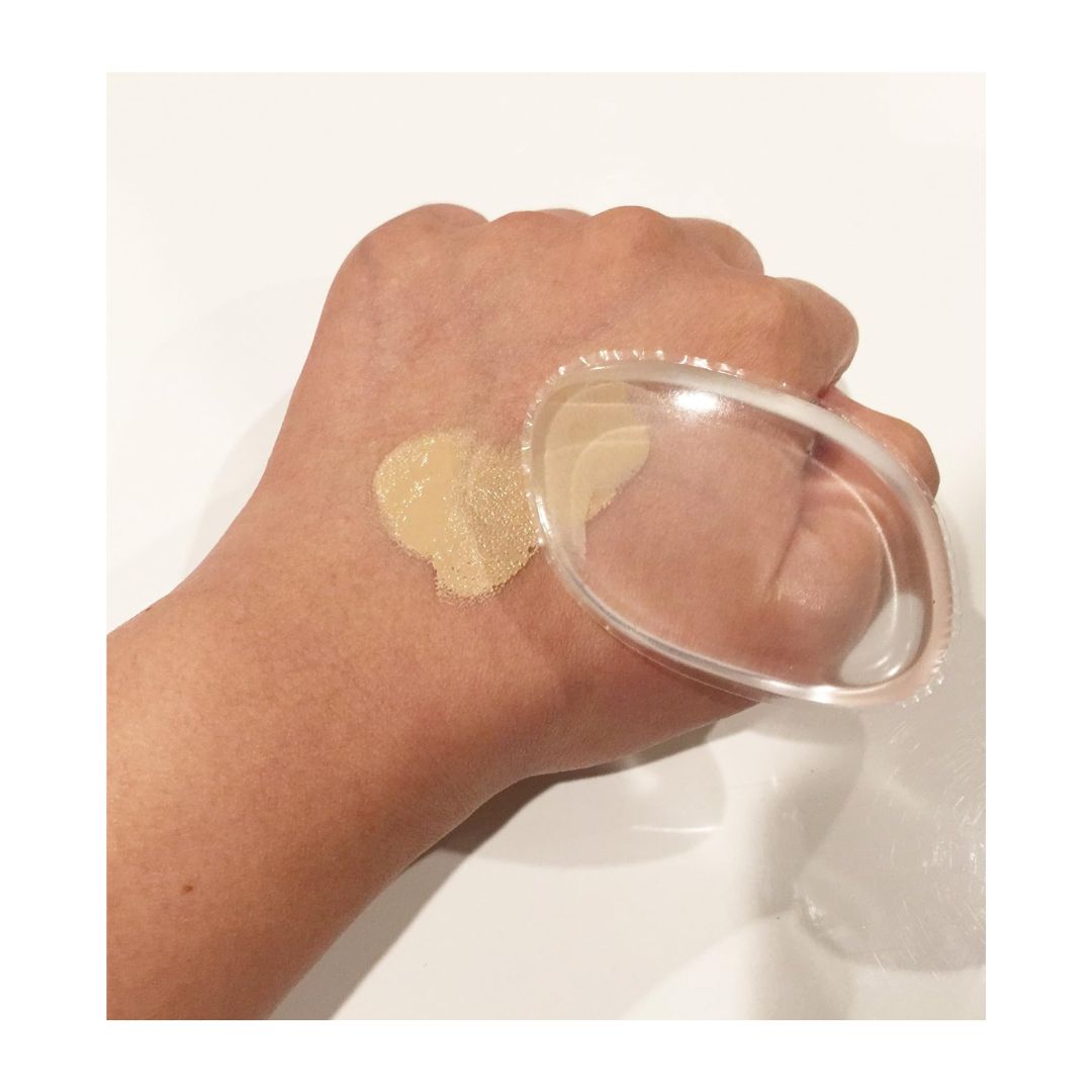 A clear, flexible makeup sponge tip being used to apply a beige liquid foundation swatch onto the back of a person's hand, set against a clean white background.