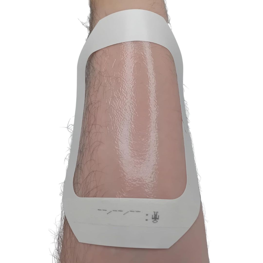 A close up of a tegaderm dressing semi applied to skin, still waiting for top layer to be removed, shown attached to a leg on a white background