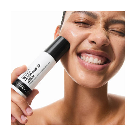 A close-up of a woman holding a white and black Hminer Labs Centon 900 Facial Barrier Serum against her cheek, with a clean white background.