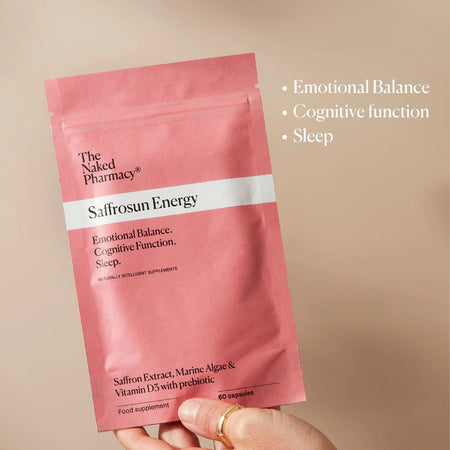A hand holds a pink stand-up pouch of The Naked Pharmacy Saffrosun Energy food supplement against a warm beige background. White text on the right lists benefits: Emotional Balance, Cognitive function, and Sleep.