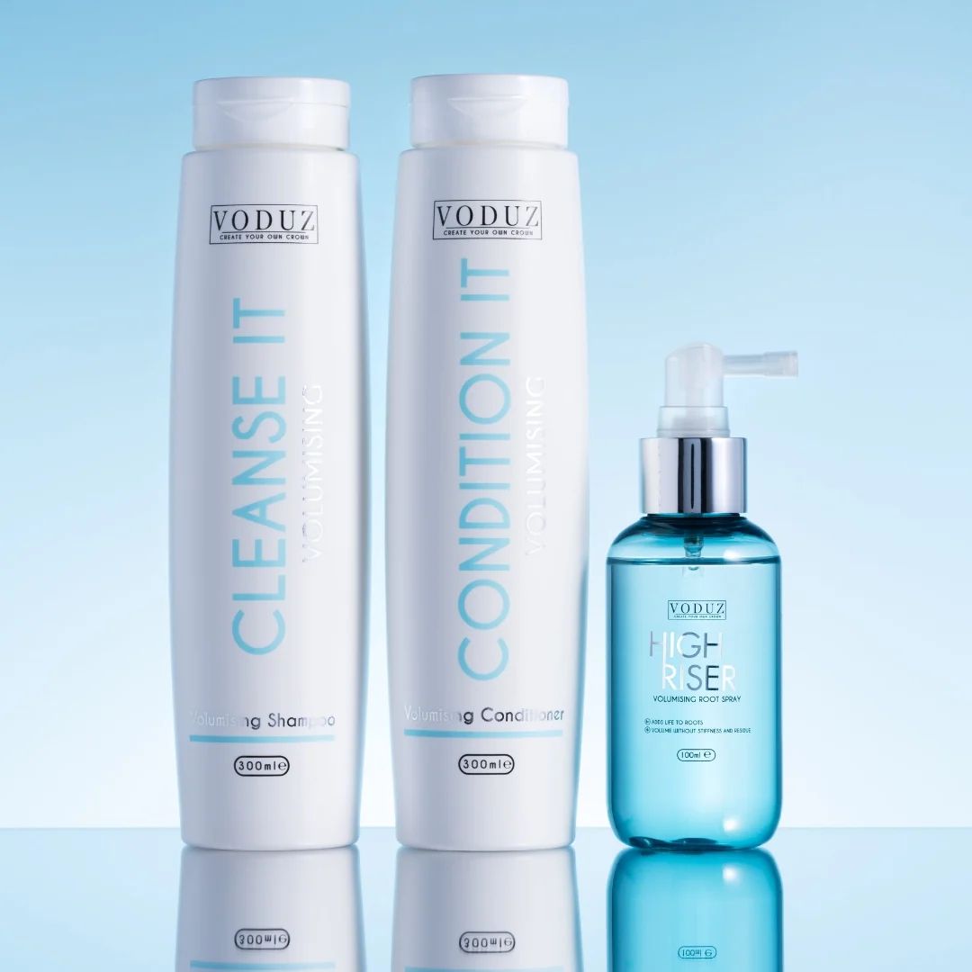 VODUZ hair care set featuring a white Cleansse It shampoo bottle, a white Condition It hair reconstructor bottle, and a blue High-Rise volumizing mist pump bottle, displayed on a reflective surface against a light blue gradient background.