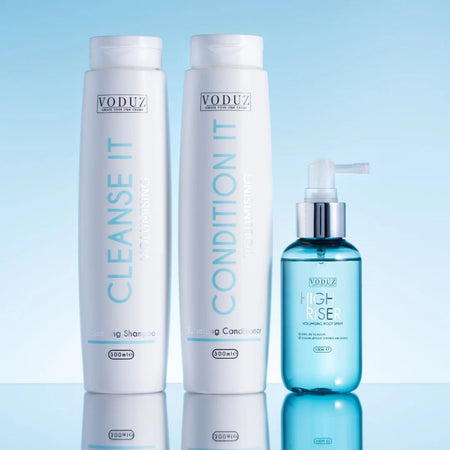 VODUZ hair care set featuring a white Cleansse It shampoo bottle, a white Condition It hair reconstructor bottle, and a blue High-Rise volumizing mist pump bottle, displayed on a reflective surface against a light blue gradient background.