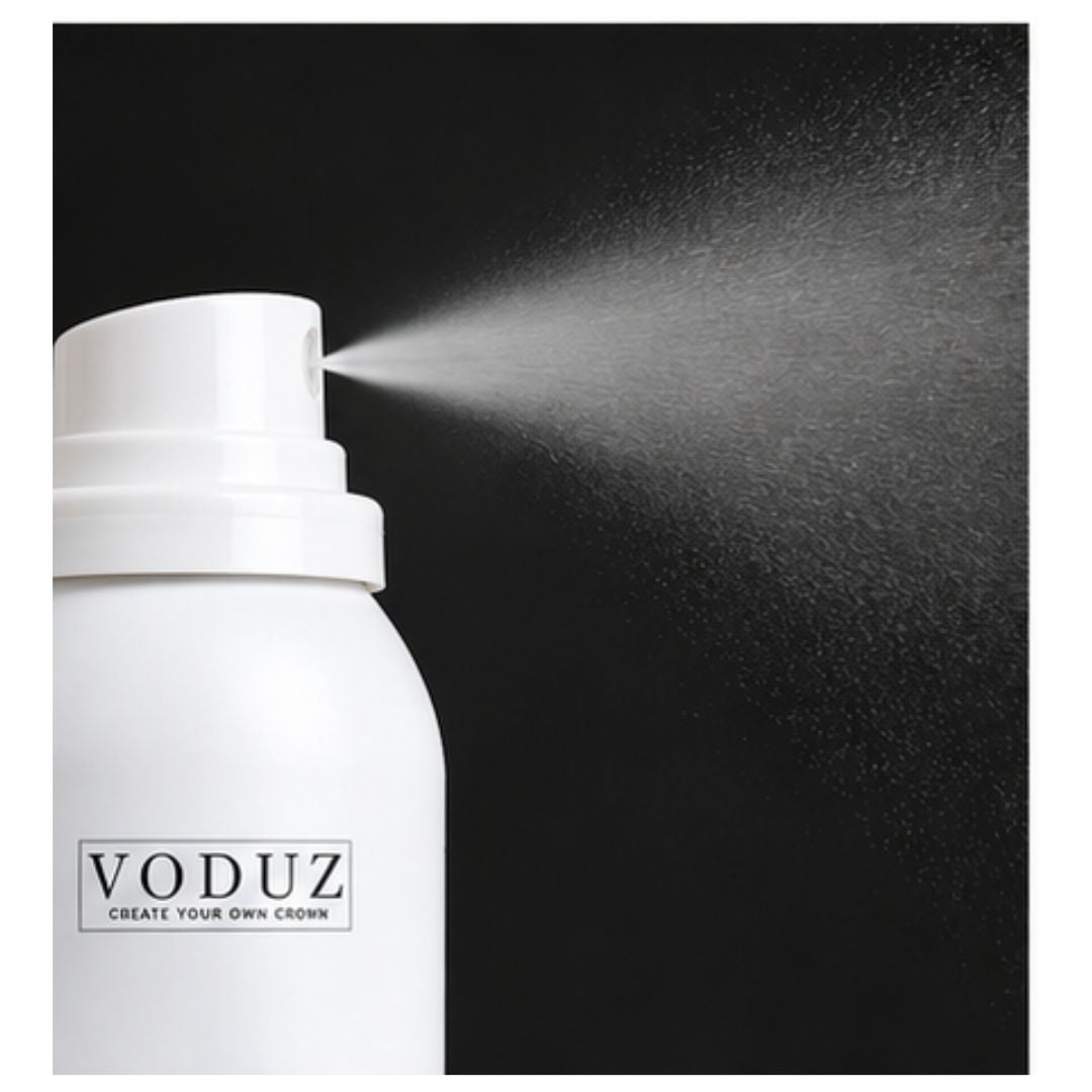 A white VODUZ hair spray bottle shown on a black background with the spray nozzle activated and mist spraying to the right.