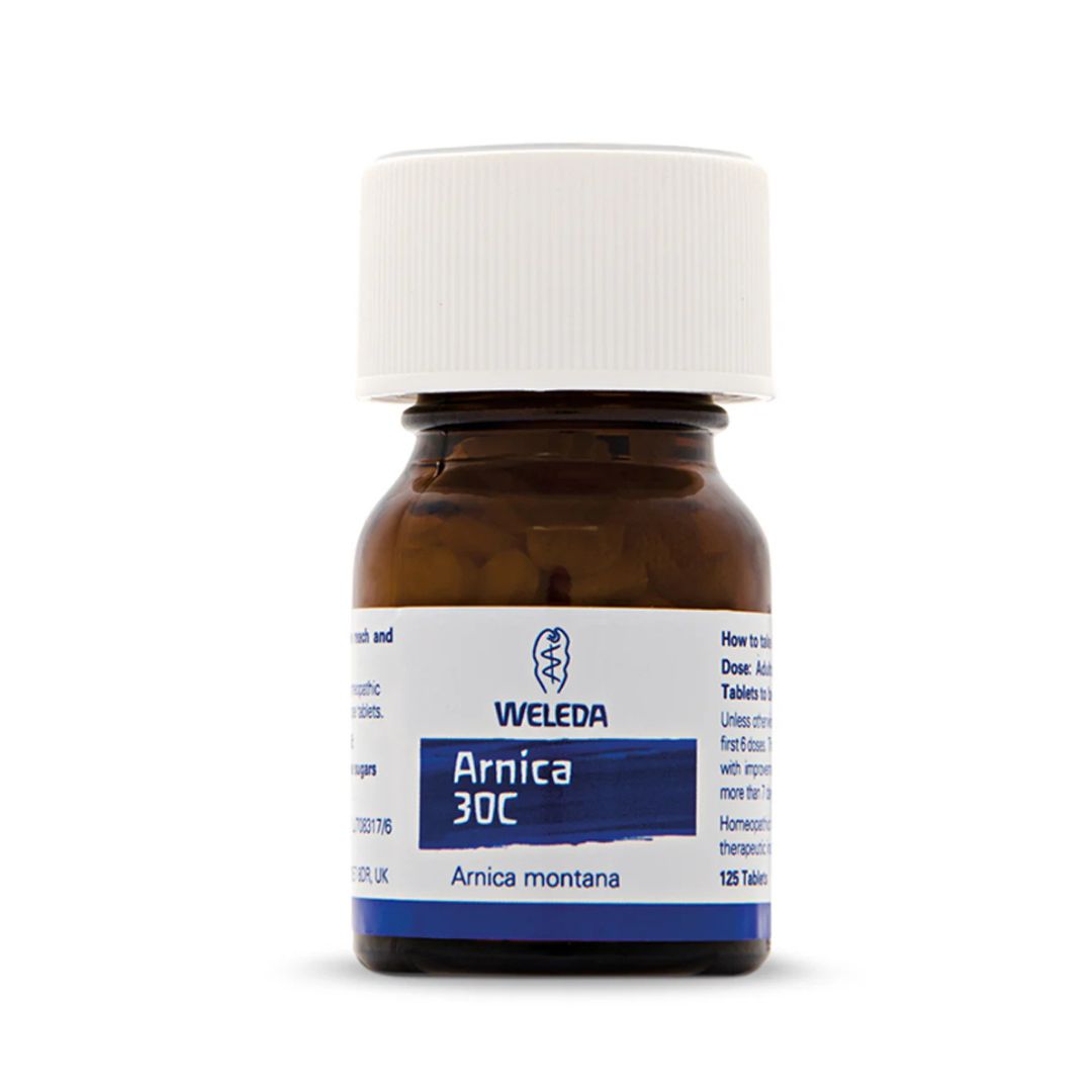 A small amber glass bottle of Weleda Arnica 30C tablets with a white screw cap and a white label featuring blue text and branding, shown on a clean white background.