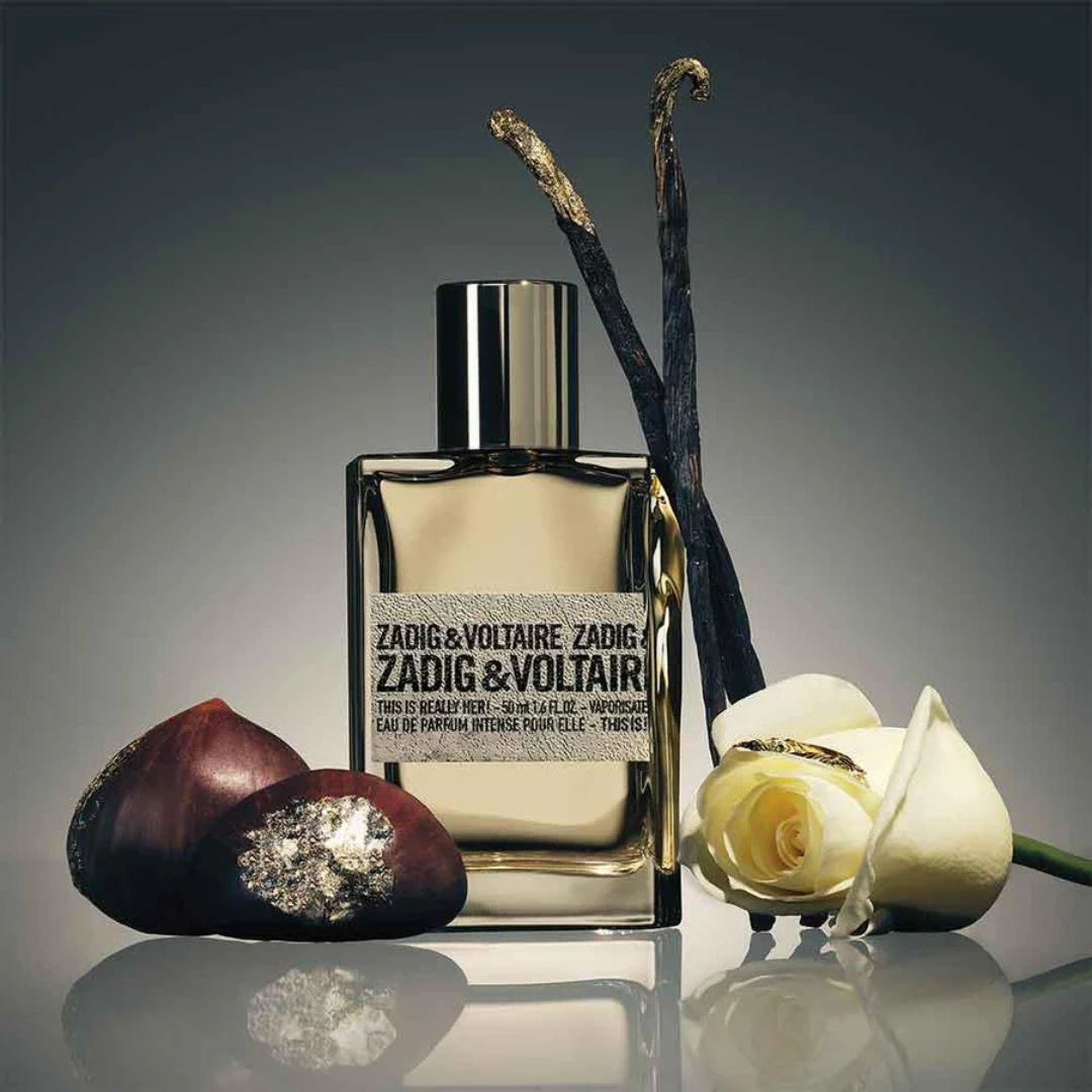 Zadig & Voltaire perfume bottle with vanilla beans, chestnuts, and a rose on a reflective surface.