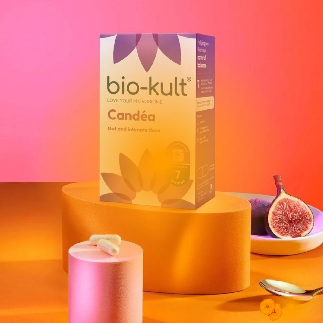 A box of bio-kult Candéa vaginal suppositories displayed on a yellow pedestal, with a pink cylindrical container, a halved fig, and a spoon on a pink and orange gradient background.