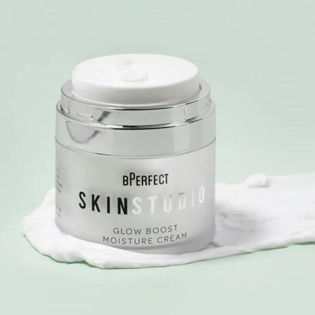 A silver cosmetic jar of bPerfect Skinstudio Glow Boost Moisture Cream with a white lid, set on a white smear of cream against a pale mint green background.