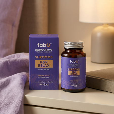 A bottle and box of fabür Shrooms R&R Relax capsules placed on a bedside table next to a lamp, with a purple blanket in the foreground.