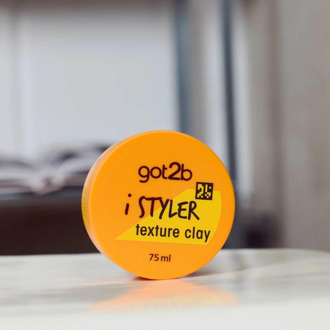 A round orange jar of got2b i STYLER texture clay sits on a white surface with a blurred indoor background.