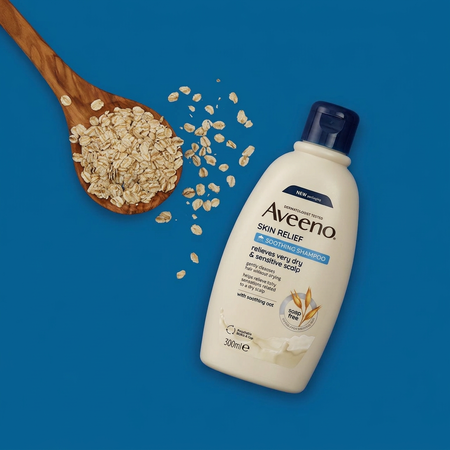 A 300ml bottle of Aveeno Skin Relief A Drier & Sensitive Scalp shampoo lies on a solid blue background next to a wooden spoon filled with rolled oats.