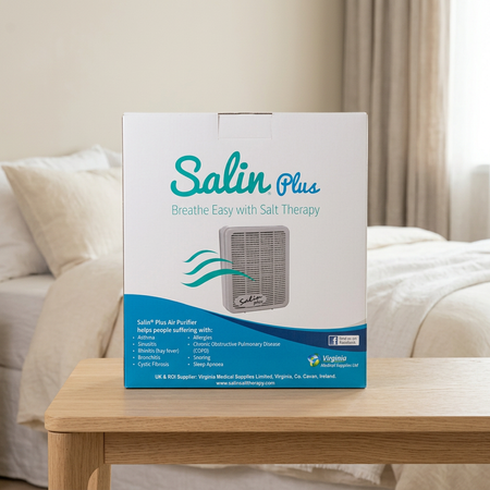 A Salin Plus salt therapy box standing on a light wood table in front of a softly blurred bedroom background with a bed and curtains.